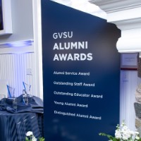 Blue sign listing the type of alumni awards with awards displayed on table next to sign